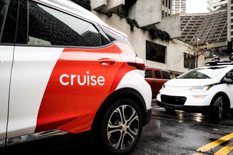 White and red Cruise autonomous car on San Francisco street with concrete Brutalist-style building in background on grey, rainy day.